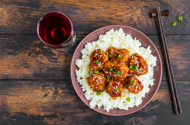 Crispy sesame chicken, chopped breast fillets, with a sticky sweet Asian sauce and white boiled rice on a plate on wooden table, top view. Crispy sesame chicken, chopped breast fillets, with a sticky sweet Asian sauce and white boiled rice on a plate on wooden table, top view. sticky sesame chicken sauces stock pictures, royalty-free photos & images