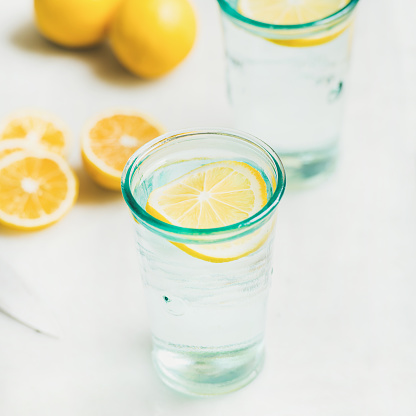 Morning detox lemon water in glasses served with fresh lemons over light grey marble background, selective focus, square crop. Clean eating, weight loss, healthy, detox, dieting concept