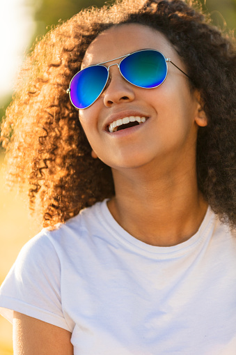 A beautiful and happy mixed race African American female girl child teenager young woman in sunshine wearing blue reflective aviator sunglasses and smiling with perfect teeth