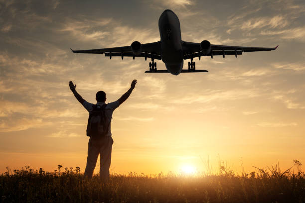 avion et silhouette d’un homme heureux debout avec les bras soulevé en place sur au coucher du soleil. paysage avec atterrissage avion de passagers voler dans le ciel jaune avec nuages, soleil et jeune homme sportif. avion - leaving business landscape men photos et images de collection