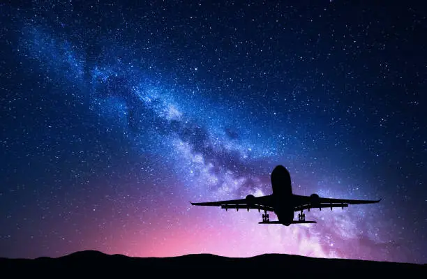 Photo of Milky Way and silhouette of a airplane. Landscape with passenger airplane is flying in the starry sky at night. Space background. Landing airliner on the background of colorful Milky Way. Aircraft