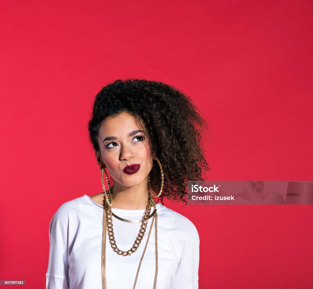 Young woman in hip-hop style against red background Studio portrait of pensive hip-hop style young latin woman wearing gold jewellery, standing against red background. Women Stock Photo