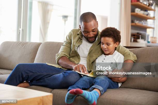 Father And Son Sitting On Sofa In Lounge Reading Book Together Stock Photo - Download Image Now