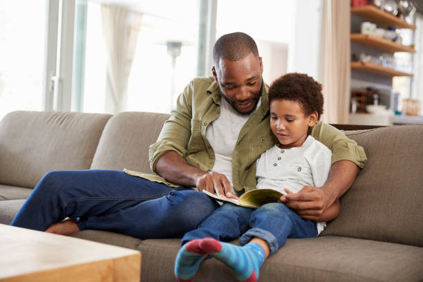 отец и сын, сидя на диване в lounge чтение книги вместе - family reading book child стоковые фото и изображения
