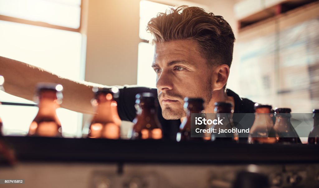 Man examining the beer bottles on conveyor at brewery factory Shot of young man examining the beer bottles on conveyor at brewery factory. Male brewer supervising the process of beer manufacturing in factory. Bottling Plant Stock Photo