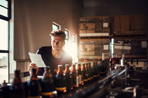 Young man supervising the process of beer manufacturing on brewery factory. Factory worker checking the process on the production line in brewery.