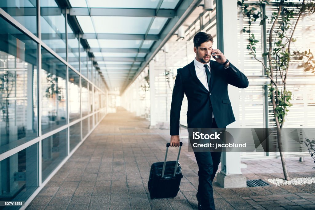 Travelling businessman making phone call Handsome young man on business trip walking with his luggage and talking on cellphone at airport. Travelling businessman making phone call. Business Travel Stock Photo