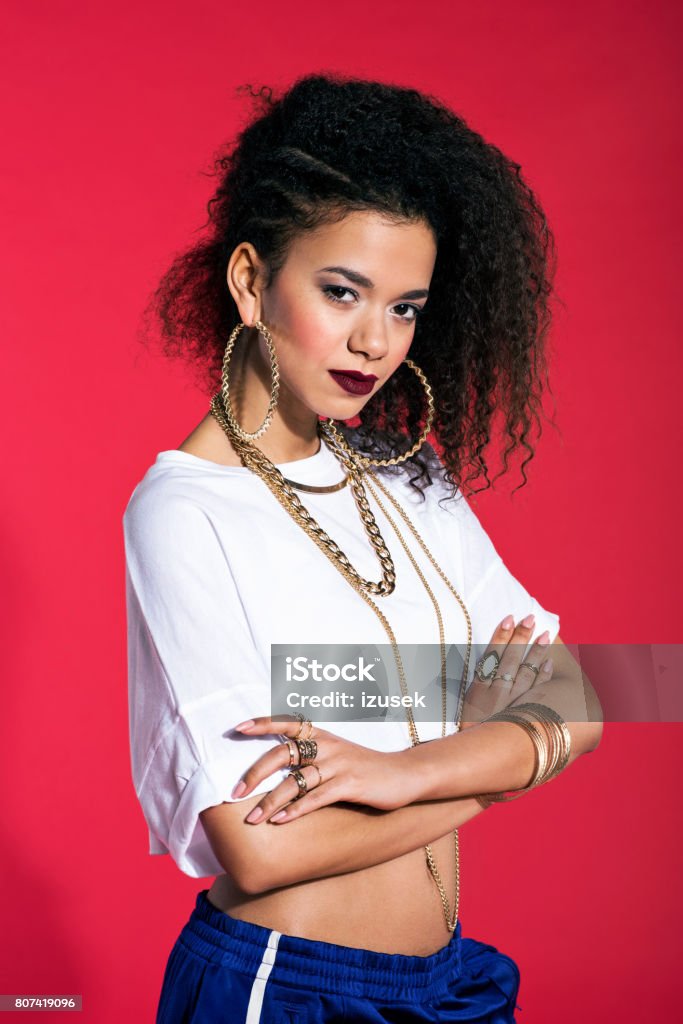 Confident latin young woman in hip-hop style against red background Studio portrait of hip-hop style young confident latin woman wearing gold jewellery and tracksuit pants. Red background. Tracksuit Stock Photo