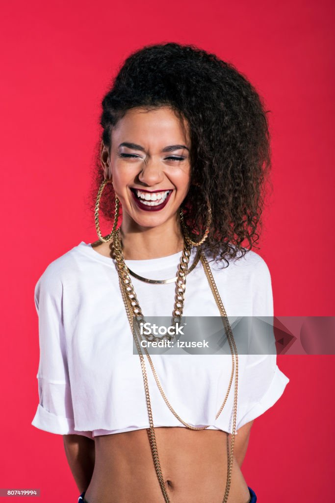 Happy young woman in hip-hop style against red background Vertical portrait of happy hip-hop style young latin woman wearing gold jewellery, laughing against red background. Make-Up Stock Photo