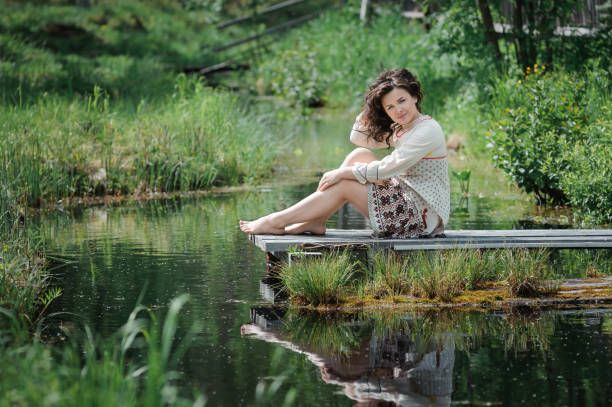 Portrait of a beautiful girl in Slavic clothes. Young barefoot woman is sitting by the pond. Summertime