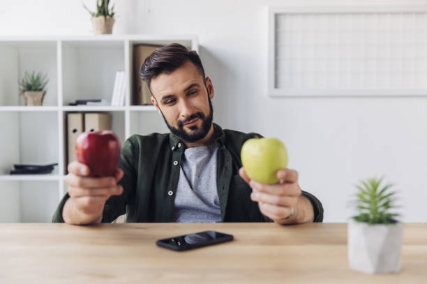 istock Smiling bearded businessman choosing between green and red apples while sitting at office table 807442550