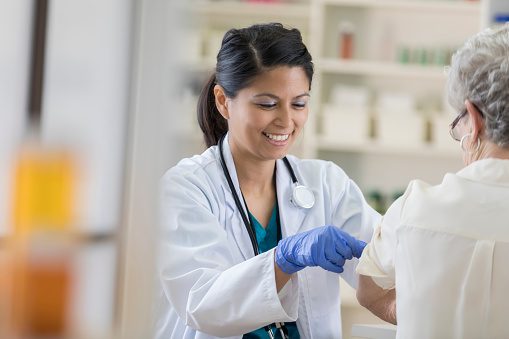 A smiling young pharmacist dons a stethoscope and completes her senior customer's flu shot by placing a band-aid on her upper arm.  She is wearing medical gloves and a lab coat.  There are shelves in the background.