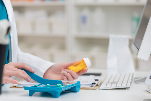 Focus is on an unrecognizable pharmacy employee as she wears scrubs and a lab coat and counts tablets to fill a prescription with one hand while holding a prescription bottle in the other.  There is a clipboard, paperwork, computer and bag on the table and shelves in the background.