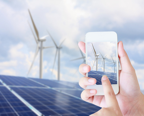 hand holding mobile phone with solar panels and wind turbines background, renewable energy