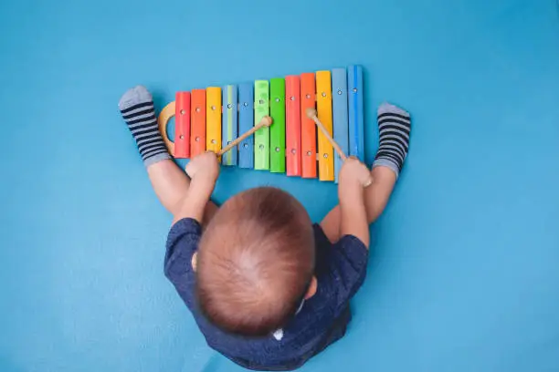 Photo of Bird eye view of Cute little Asian 18 months / 1 year old baby boy child hold sticks & plays a musical instrument colorful wooden toy xylophone