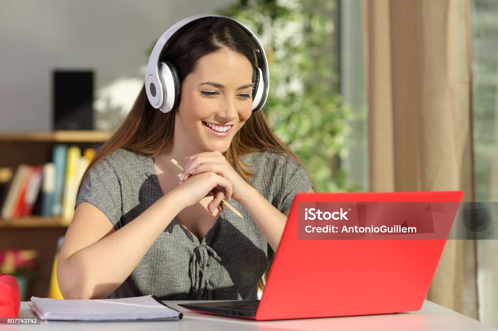 Student watching video tutorials on line Portrait of a beautiful student viewing and listening video tutorials on line with headphones and a red pc sitting in a table at home E-Learning Stock Photo