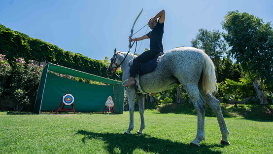 Young man is enjoying horseback shooting archery in nature in grass.