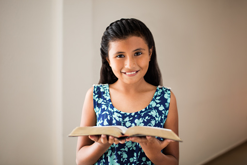 A happy latin girl standing, holding an open Bible and smiling at the camera.