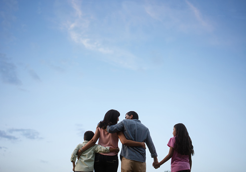 Back view of a latin family of four standing outdoors, embracing and holding hands with the sky in the background.