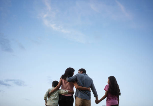 Latin family standing together with sky in background Back view of a latin family of four standing outdoors, embracing and holding hands with the sky in the background. family holding hands stock pictures, royalty-free photos & images