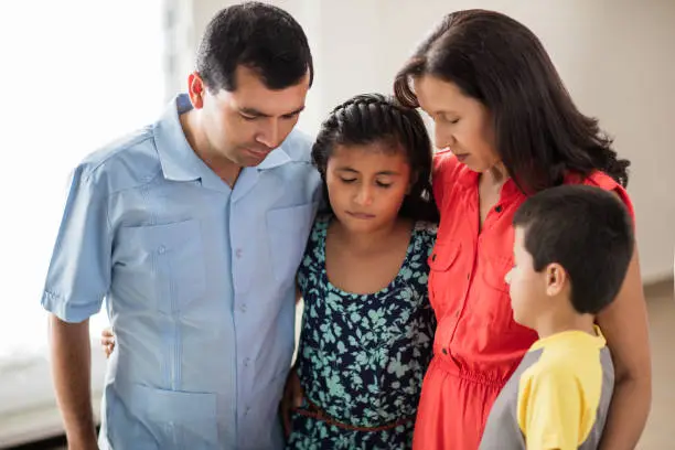 A latin family with two children standing, embracing and praying with their eyes closed.