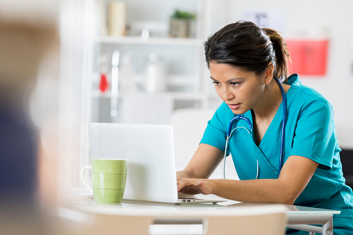 Serious mid adult female healthcare professional uses a laptop in her office. She is wearing scrubs and a stethoscope.