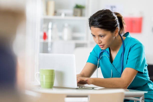 Female healthcare professional uses a laptop Serious mid adult female healthcare professional uses a laptop in her office. She is wearing scrubs and a stethoscope. doctor utensils stock pictures, royalty-free photos & images