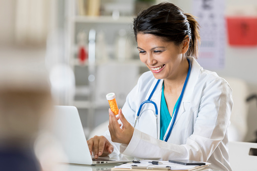 Smiling female physician reviews a patient's medication. She is also using a laptop to do research on the medication.