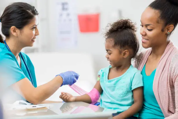 Cheerful female phlebotomist wraps a preschool age girl's arm with a pink bandage after drawing blood from the girl's arm. The girl is sitting in her mom's lap.