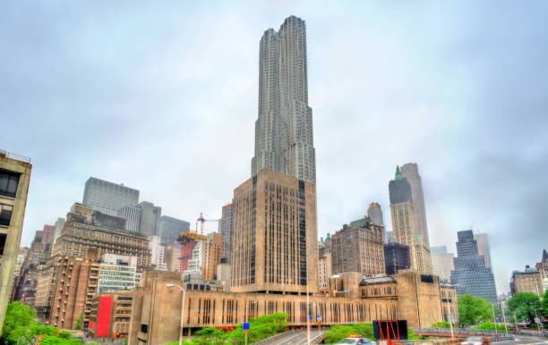 A street-level view in New York City showcases the contrast between historic architecture and sleek modern skyscrapers. Ornate stonework and Gothic details with a golden spire in the foreground rise alongside a row of early 20th-century facades, while a contemporary glass tower dominates the background against a clear blue sky.