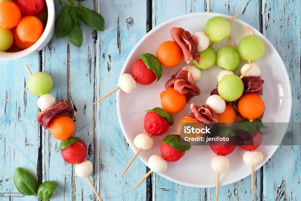 Plate of summer fruit skewers on a rustic blue wood background Plate of delicious summer fruit skewers with melon, cheese and prosciutto on a rustic blue wood background Skewer Stock Photo