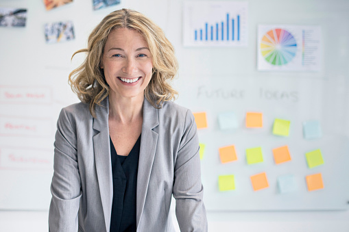 Portrait of confident businesswoman in office. Smiling female professional is standing against whiteboard with charts and adhesive notes. Smiling manager is in businesswear at workplace.