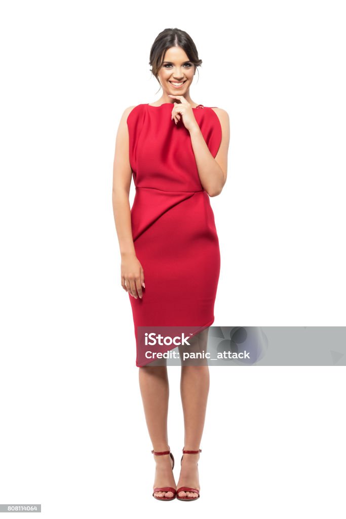 Shy timid young woman in red elegant dress smiling at camera with hand under chin Shy timid young woman in red elegant dress smiling at camera with hand under chin. Full body length portrait isolated on white studio background. Women Stock Photo