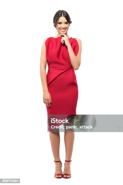 Shy Timid Young Woman In Red Elegant Dress Smiling At Camera With Hand Under Chin Stock Photo - Download Image Now