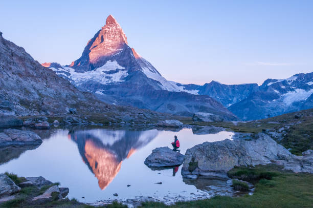 S Early morning dawn scene of sunrise on the Matterhorn Mountain reflecting pink in the lake with male man on rock with red down jacket with clear blue sky Gornergrat Zermatt Matterhorn Europe pennine alps stock pictures, royalty-free photos & images