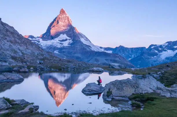 Early morning dawn scene of sunrise on the Matterhorn Mountain reflecting pink in the lake with male man on rock with red down jacket with clear blue sky Gornergrat Zermatt Matterhorn Europe