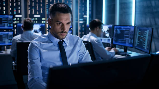 istock Close-up of Technical Engineer Working on His Computer With Multiple Displays in Monitoring Room. In the Background His Colleagues with Graphics and Various Data on Their Monitors. 808155072