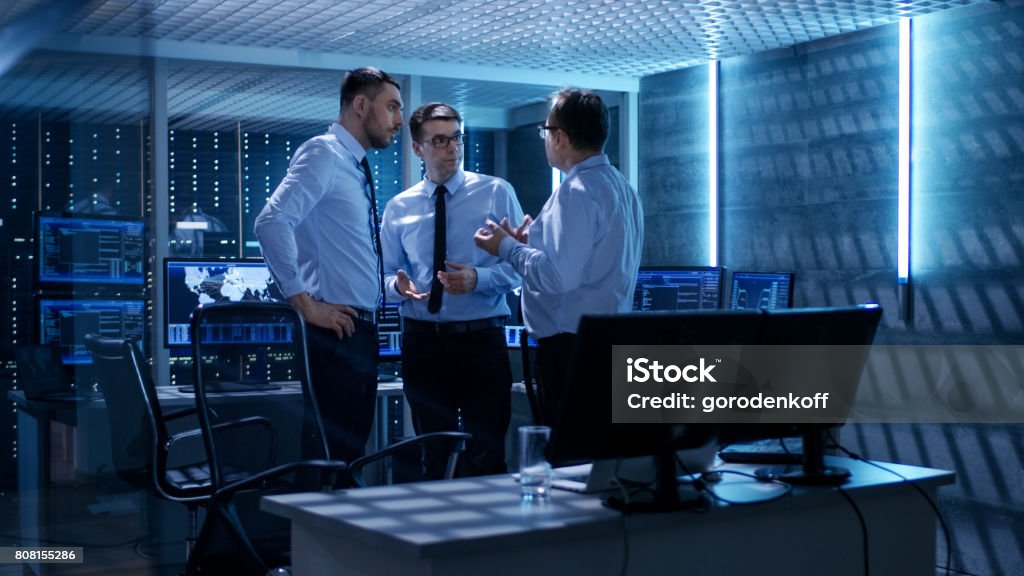 Team of Technical Moderators Have Active Discussion in Monitoring Room. System Control Room is full of Working Displays Showing Various Data and Has Servers Racks. Advice Stock Photo