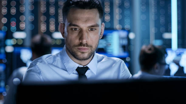 Close-up of a Professional Technical Controller Sitting at His Desk with Multiple Displays Before Him. In the Background His Colleagues Working in System Control Center. Close-up of a Professional Technical Controller Sitting at His Desk with Multiple Displays Before Him. In the Background His Colleagues Working in System Control Center. facilities protection services stock pictures, royalty-free photos & images