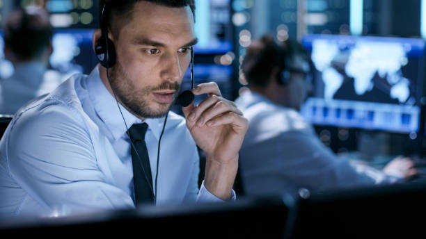 In Monitoring Room Technical Support Specialist Speaks into Headset. His Colleagues are Working in the Background. In Monitoring Room Technical Support Specialist Speaks into Headset. His Colleagues are Working in the Background. facilities protection services stock pictures, royalty-free photos & images