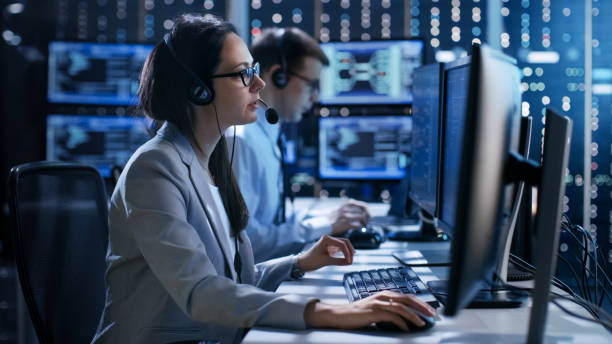 la donna che lavora in un team di supporto tecnico fornisce istruzioni con l'aiuto delle cuffie. in background le persone che lavorano e monitora mostrano varie informazioni. - facilities protection services foto e immagini stock