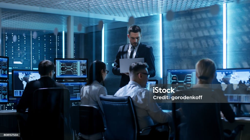 Professional IT Engineers Working in System Control Center Full of Monitors and Servers. Supervisor Holds Laptop and Holds a Briefing. Possibly Government Agency Conducts Investigation. IT Support Stock Photo