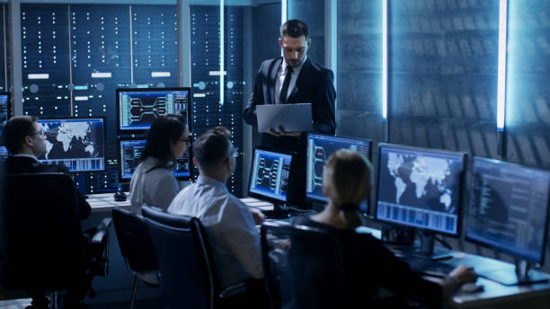 profesional ingenieros trabajando en el centro de control de sistema completo de monitores y servidores. supervisor tiene laptop y lleva a cabo una reunión informativa. posiblemente la agencia gubernamental realiza investigación. - servicio federal de protección fotografías e imágenes de stock