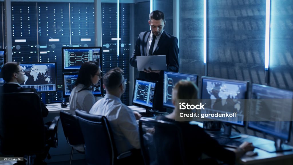 Professional IT Engineers Working in System Control Center Full of Monitors and Servers. Supervisor Holds Laptop and Holds a Briefing. Possibly Government Agency Conducts Investigation. Cross Section Stock Photo