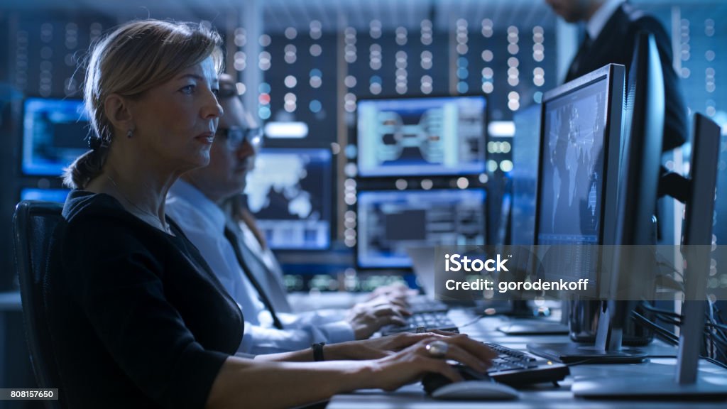 Female Government Employee Works in a Monitoring Room. In The Background Supervisor Holds Briefing. Possibly Government Agency Conducts Investigation. Network Security Stock Photo