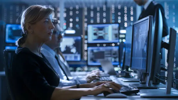 Photo of Female Government Employee Works in a Monitoring Room. In The Background Supervisor Holds Briefing. Possibly Government Agency Conducts Investigation.