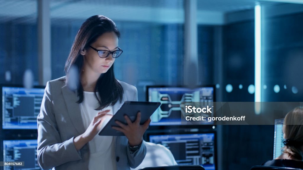 Young Female Government Employee Wearing Glasses Uses Tablet in System Control Center. In the Background Her Coworkers are at Their Workspaces with many Displays Showing Valuable Data. Network Security Stock Photo