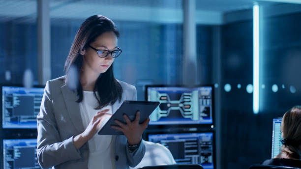 Young Female Government Employee Wearing Glasses Uses Tablet in System Control Center. In the Background Her Coworkers are at Their Workspaces with many Displays Showing Valuable Data. Young Female Government Employee Wearing Glasses Uses Tablet in System Control Center. In the Background Her Coworkers are at Their Workspaces with many Displays Showing Valuable Data. cybersecurity stock pictures, royalty-free photos & images