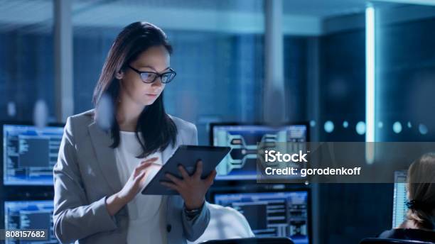 Young Female Government Employee Wearing Glasses Uses Tablet In System Control Center In The Background Her Coworkers Are At Their Workspaces With Many Displays Showing Valuable Data Stock Photo - Download Image Now