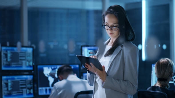 la giovane ingegnere femminile utilizza il tablet nel centro di controllo del sistema. in background i suoi colleghi si trovano nelle aree di lavoro con molti display che mostrano dati preziosi. - facilities protection services foto e immagini stock